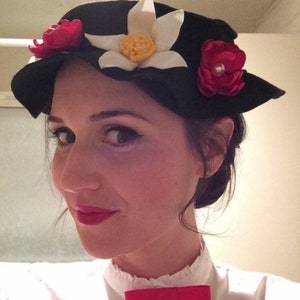 May include: A woman wearing a black hat with white and red flowers. She is wearing a white shirt with a red bow tie.