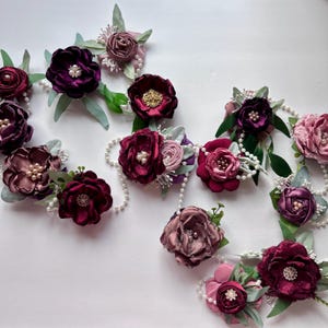 Puede incluir: Una variedad de accesorios florales hechos a mano en tonos burdeos, ciruela y malva. Cada flor está adornada con perlas y vegetación, algunas unidas a hilos de perlas. Adecuado para ramilletes o accesorios para el cabello.
