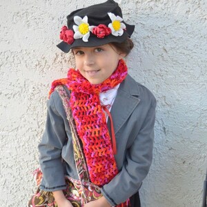 May include: A young person dressed as a fictional character wearing a black hat with red and white flowers, a red and pink crocheted scarf, and a gray jacket.