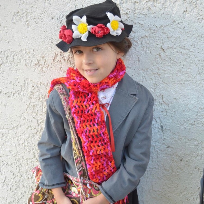 May include: A young person dressed as a fictional character wearing a black hat with red and white flowers, a red and pink crocheted scarf, and a gray jacket.