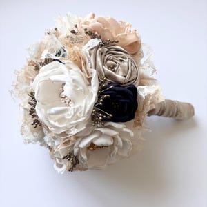 Puede incluir: Un ramo de novia artesanal con flores de tela en marfil, rosa y azul marino. El ramo incluye encaje y detalles de cuentas, con un mango envuelto. El arreglo muestra una variedad de texturas y colores, ideal para una boda.