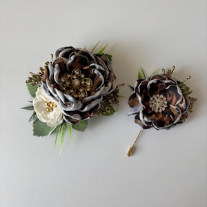 Puede incluir: Un conjunto de dos broches florales con un tejido de estampado de leopardo marrón y blanco con un centro de pedrería. El broche más grande tiene una flor blanca con detalles dorados y el broche más pequeño es un alfiler.