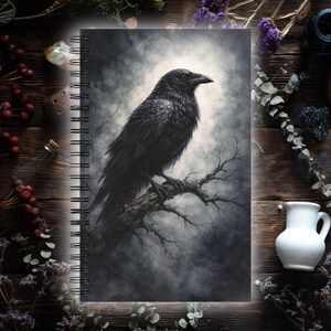Puede incluir: Cuaderno con espiral que presenta una ilustración detallada de un cuervo negro posado en una rama desnuda sobre un fondo gris brumoso. El diseño evoca un tema gótico o de fantasía.