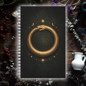 Könnte beinhalten: Ein Spiralnotizbuch mit einem schwarzen, ledertexturierten Einband. Der Einband zeigt ein goldenes Ouroboros-Design mit Sternen und Monden. Das Notizbuch ist von getrockneten Blumen und anderen dekorativen Gegenständen umgeben.