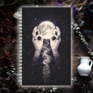 Puede incluir: Cuaderno con espiral con un diseño celestial. La portada muestra manos sosteniendo una luna llena brillante contra un cielo estrellado. Un manojo de salvia con humo ascendente está debajo de la luna. El cuaderno tiene un fondo oscuro y encuadernación en espiral plateada.