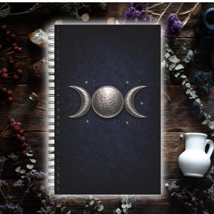 Può includere: Un taccuino a spirale nero con una copertina con un design a tripla luna argentata. Il design presenta un cerchio centrale e mezzelune, su uno sfondo blu scuro con piccole stelle bianche. Il taccuino è circondato da elementi in legno.