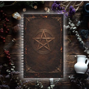 Op de afbeelding: Een bruin spiraalgebonden notitieboek met een pentagramontwerp. De kaft heeft een getextureerde, leerachtige uitstraling met decoratieve hoekaccenten. Het notitieboek is omgeven door gedroogde bloemen, kristallen en andere mystieke items op een houten oppervlak.