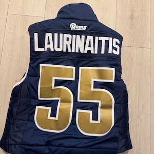 May include: A navy blue quilted vest with the name "LAURINAITIS" in white lettering and the number "55" in gold. The vest has a "Rams" logo at the top. The vest is lying on a light-colored wooden surface.