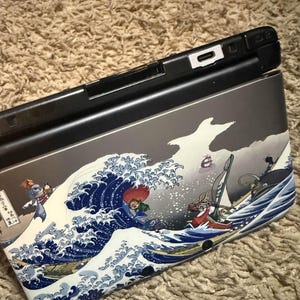 May include: A black handheld gaming console with a custom case featuring a stylized wave design in shades of blue, white, and gray. The artwork includes figures in boats and a red-haired character.