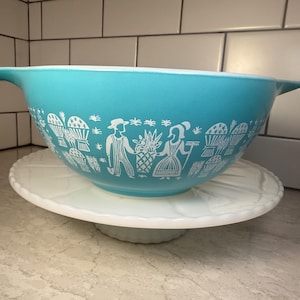 May include: A turquoise mixing bowl with white handles and a white graphic print featuring figures of a person with a shovel and a person with a rake, surrounded by wheat sheaves and baskets, resting on a white scalloped glass cake stand.