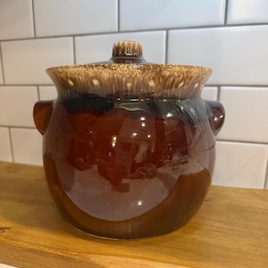 May include: A vintage brown ceramic bean pot with a lid. The pot has a glossy finish with a darker brown base and lighter brown rim. It features two side handles and a small knob on the lid. The pot is sitting on a wooden surface.