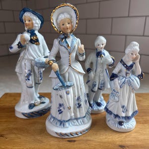 May include: A set of four porcelain figurines featuring two individuals in period attire with blue and gold accents. The figures are dressed in white coats, dresses, and hats, with intricate blue floral patterns and gold trim on their clothing.