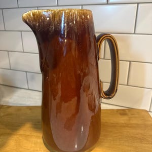 May include: A brown ceramic pitcher with a glossy finish. The pitcher has a dark brown handle and a spout. The top edge has a lighter brown, textured design. The pitcher is sitting on a wooden surface.