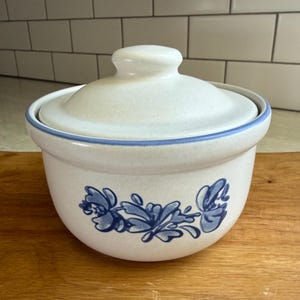 May include: A round, off-white ceramic casserole dish with a matching lid. The exterior features a blue floral pattern and a thin blue rim around the top edge. The surface has a speckled texture, and the lid includes a central knob handle.