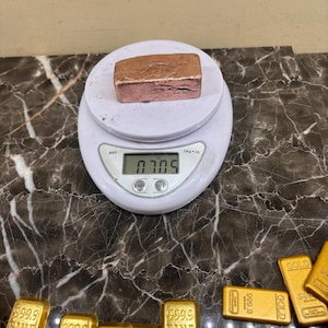 May include: A copper bar weighing 0.705 ounces is displayed on a digital scale. Several gold bars are visible below the scale. The gold bars are labeled with the text "999.9 FINE GOLD".