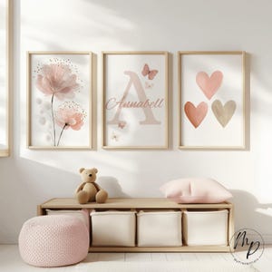 Könnte beinhalten: Drei gerahmte Kunstdrucke mit einem Blumendesign, einem Buchstaben "A" mit dem Namen "Annabell" und drei Herzen. Die Drucke sind in einem hellrosa und beigen Farbschema gehalten. Eine Holzbank mit Aufbewahrungswürfeln, ein Teddybär und ein rosa Hocker befinden sich im Vordergrund.