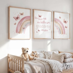 May include: Three framed art prints with a watercolor rainbow and butterflies in soft pink and beige tones. The middle print features the German text "Du bist nie zu klein um großartig zu sein". A wooden bed with teddy bears and pillows is in the foreground.