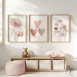 May include: Three framed art prints featuring floral and heart designs in soft pink and neutral tones. The prints are displayed above a wooden storage bench with a teddy bear, a pink cushion, and a pink knitted pouffe.
