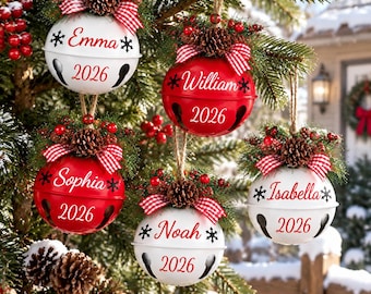 Può includere: Cinque decorazioni natalizie a forma di campanello rotondo in rosso e bianco, personalizzate con un nome e l'anno 2026 in rosso. Ogni ornamento è sormontato da una pigna, bacche rosse e un fiocco a quadretti rossi e bianchi.