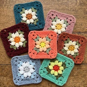 Op de afbeelding: Een verzameling van zeven gehaakte vierkante onderzetters in verschillende kleuren. Elke onderzetter heeft een centraal bloemmotief met witte bloemblaadjes en een contrasterend centrum, omgeven door een vierkante rand. Kleuren: teal, roze, bordeaux, oranje, lichtblauw en groen.