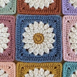 May include: A collection of square crochet motifs arranged in a grid. Each square features a central daisy pattern with a mustard yellow center and white petals, framed by a solid-colored border in shades of blue, pink, green, and brown.