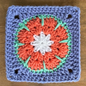 May include: A square crochet motif featuring a central white flower, surrounded by layers of coral-orange petals, accented with light green stitches, and framed by a light purple border with four corner spaces, creating a geometric floral design.