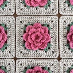 May include: A collection of square crochet motifs arranged in a grid pattern. Each square features a three-dimensional pink flower with layered petals at the center, surrounded by small green leaves, all set against a textured beige yarn background.