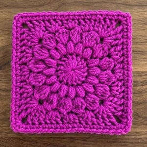 Op de afbeelding: Een vierkant, magenta gehaakt stuk met een bloemmotief. Het midden heeft een cirkelvormig patroon, omringd door bloembladachtige vormen. De randen zijn netjes afgewerkt, waardoor een getextureerde, handgemaakte uitstraling ontstaat. Het stuk is waarschijnlijk een decoratief element of onderdeel van een groter knutselproject.