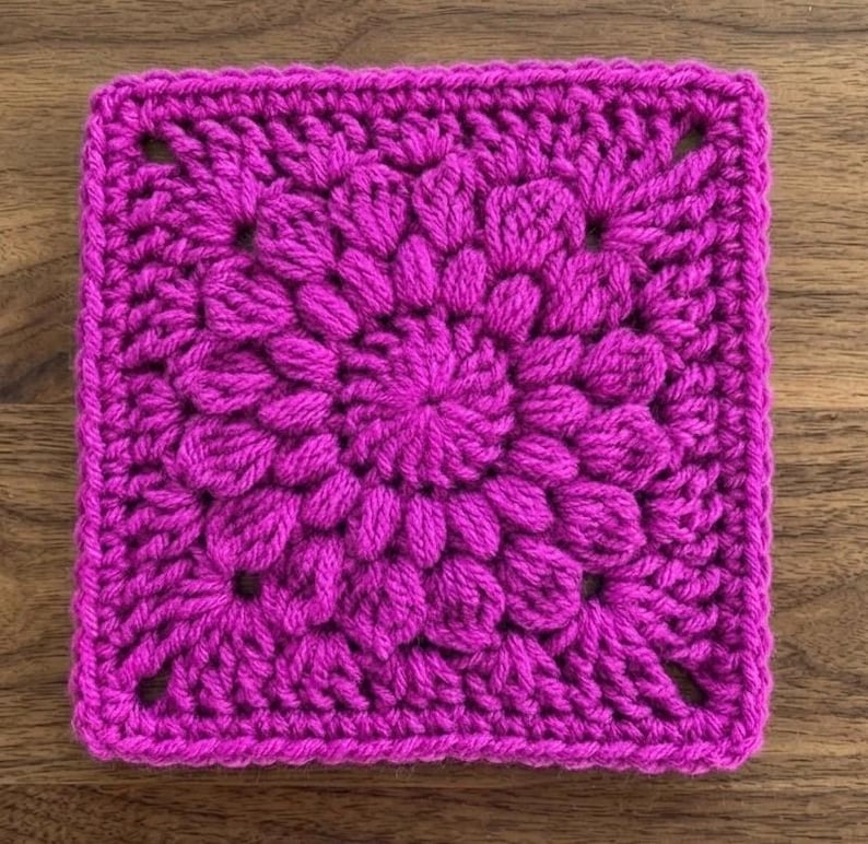 Op de afbeelding: Een vierkant, magenta gehaakt stuk met een bloemmotief. Het midden heeft een cirkelvormig patroon, omringd door bloembladachtige vormen. De randen zijn netjes afgewerkt, waardoor een getextureerde, handgemaakte uitstraling ontstaat. Het stuk is waarschijnlijk een decoratief element of onderdeel van een groter knutselproject.