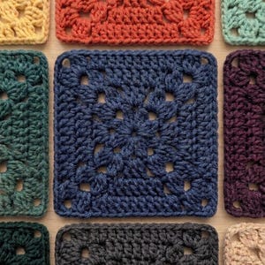 May include: Close-up of various crocheted granny squares in a variety of colors, including blue, orange, green, and purple. The squares are arranged on a light brown surface, showcasing the texture and detail of the handmade crochet work.