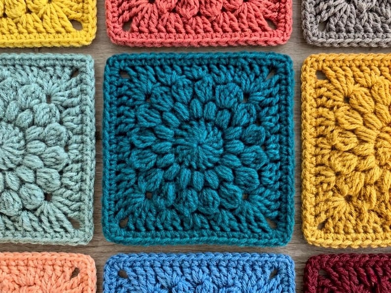 Op de afbeelding: Close-up van verschillende kleurrijke haakvierkanten in tinten van turquoise, geel, koraal en blauw. Elk vierkant heeft een uniek textuurontwerp, met een centraal bloemmotief. De vierkanten zijn dicht bij elkaar geplaatst.