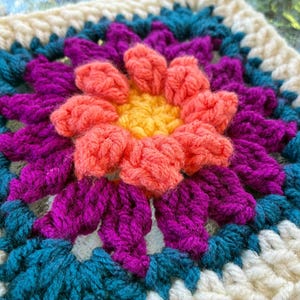 Puede incluir: Un cuadrado de ganchillo tipo granny square que presenta una flor amarilla central, rodeada por capas de pétalos de lana en color coral, magenta y azul cerceta. El borde exterior está hecho con lana color crema, creando un diseño floral texturizado.