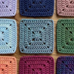 May include: A collection of colorful, square crochet granny squares in various shades, including blue, green, pink, and purple. Each square features a central design and a textured border, showcasing the intricate details of the handmade craft. The squares are arranged on a wooden surface.