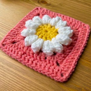 May include: A square, handmade crochet piece featuring a daisy design. The center of the flower is yellow, surrounded by white petals. The square base is a solid coral color, with a textured stitch pattern.