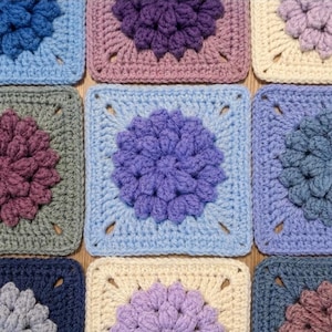 Peut inclure: Gros plan sur une collection de carrés au crochet colorés. Chaque carré présente un motif floral central dans des tons de bleu, de violet, de rose et de crème. Les carrés sont disposés en grille, mettant en valeur la texture et les détails de l'artisanat.