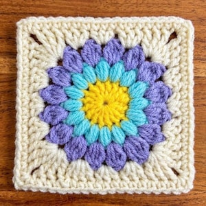 May include: A square, handmade crochet piece featuring a floral design. The center is yellow, surrounded by layers of turquoise and purple petals. The outer border is a cream color, creating a vibrant, textured textile.