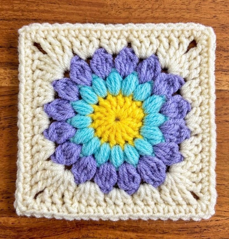 May include: A square, handmade crochet piece featuring a floral design. The center is yellow, surrounded by layers of turquoise and purple petals. The outer border is a cream color, creating a vibrant, textured textile.