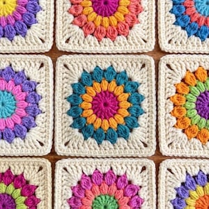 Può includere: Primo piano di nove quadrati granny crochet colorati. Ogni quadrato presenta un motivo floreale centrale in tonalità vivaci come rosa, arancione, blu e viola, circondato da un bordo color crema. I quadrati sono disposti a griglia.