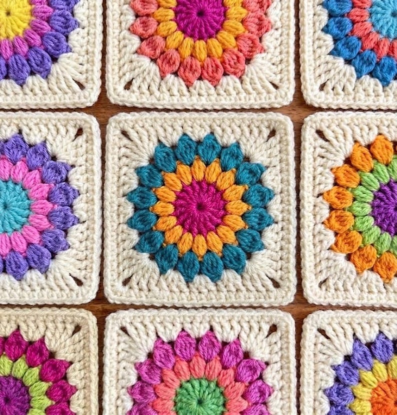 May include: Close-up of nine colorful crochet granny squares. Each square features a central flower design in vibrant hues like pink, orange, blue, and purple, surrounded by a cream-colored border. The squares are arranged in a grid pattern.