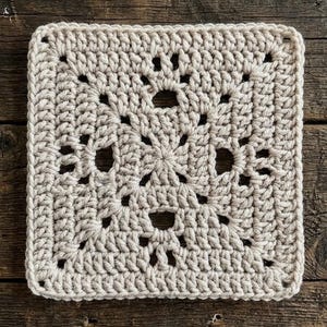 May include: A square crochet motif in a light beige yarn color. The piece features a central star-like pattern radiating outward with openwork spaces between the stitches, finished with a solid border of double crochet stitches along all four edges.