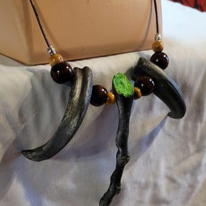 May include: A handcrafted necklace featuring dark gray claw-shaped pendants, wooden beads, and a central green and black branch-like element. The necklace is strung on a brown cord.