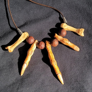 May include: A handcrafted necklace featuring bone-shaped pendants and carved wooden beads strung on a brown cord. The pendants have a natural, off-white color, while the beads are a reddish-brown. The necklace is displayed against a dark background.