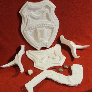 Könnte beinhalten: Weiße, unmontierte 3D-gedruckte Teile eines Schildes und anderer Komponenten, darunter zwei gebogene Hörner, ein Bein und kleine Hardware. Die Teile sind auf einer roten Oberfläche angeordnet.