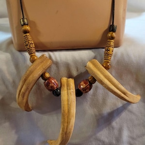 May include: A handcrafted necklace featuring large, curved, light-brown wooden pendants. The necklace includes decorative wooden beads with etched patterns and smaller beads in red, black, and brown. The necklace is strung on a dark cord.