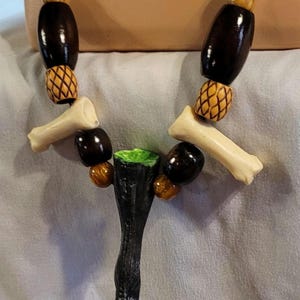 May include: A handcrafted necklace featuring a dark brown cord, bone-shaped beads, dark brown oval beads, and decorative wooden beads. The pendant is a black, stylized claw with green accents.
