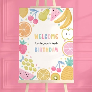 May include: A colorful birthday sign featuring watercolor illustrations of various fruits, including strawberries, bananas, and oranges. The sign reads "WELCOME to Emma's 2nd BIRTHDAY" in pastel colors, surrounded by fruit and floral accents.
