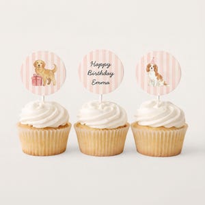 May include: Three cupcakes with white frosting and decorative toppers. The toppers feature a golden retriever with a gift, the text "Happy Birthday Emma", and a Cavalier King Charles Spaniel wearing a party hat.