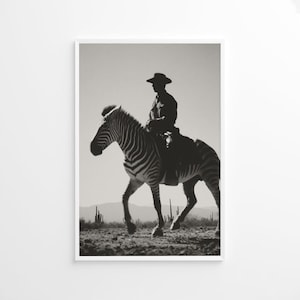 Op de afbeelding: Zwart-witfoto van een persoon die op een zebra rijdt in een woestijnlandschap. De ruiter, met hoed, is een silhouet. De zebra loopt, met cactussen en heuvels op de achtergrond.