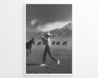 May include: Black and white framed print of a cowboy swinging a bat in a field with horses and mountains in the background. The cowboy is wearing a hat, jeans, and white shoes. The horses are dark and grazing.
