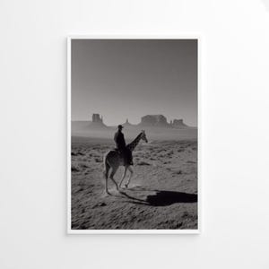 Può includere: Fotografia in bianco e nero di una persona a cavallo di una giraffa in un paesaggio desertico. La persona indossa un cappello e abiti scuri. La giraffa si muove, proiettando un'ombra. Sullo sfondo, mesas sotto un cielo velato.